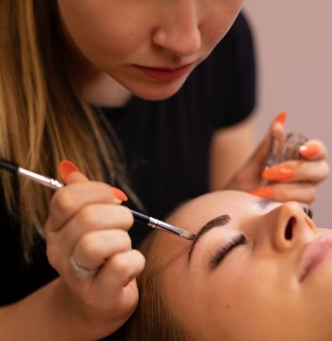 Young cosmetologist applying dye through brush while coloring female client's eyebrow at salon